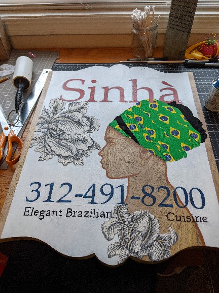 Wood sign with white background, flowers and women in profile reads: Sinha: Elegant Brazilian Cuisine.